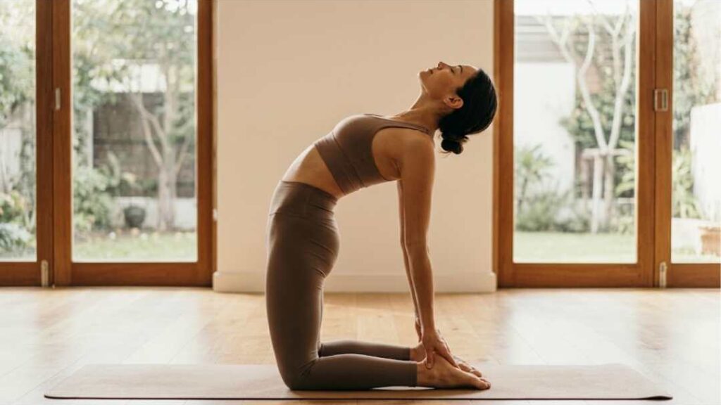 Woman performing Ustrasana (Camel Pose) yoga posture on a mat indoors, stretching the neck and chest for improved thyroid health.